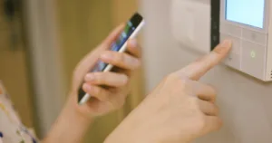 Person holding a smartphone while pressing a button on a smart home control panel mounted on a wall.