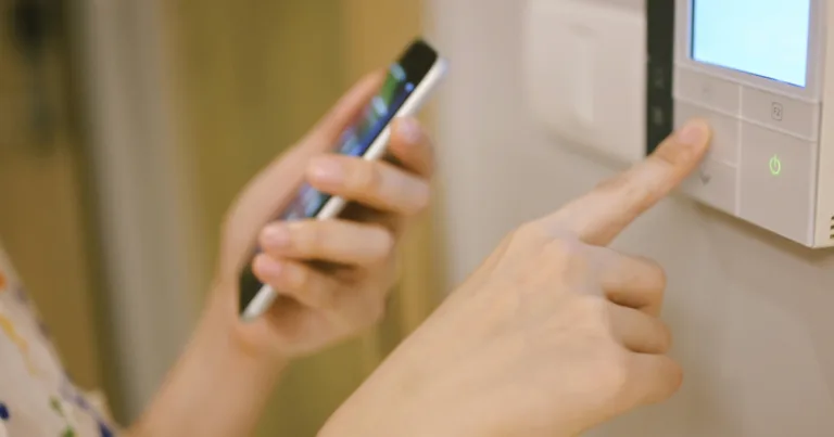 Person holding a smartphone while pressing a button on a smart home control panel mounted on a wall.