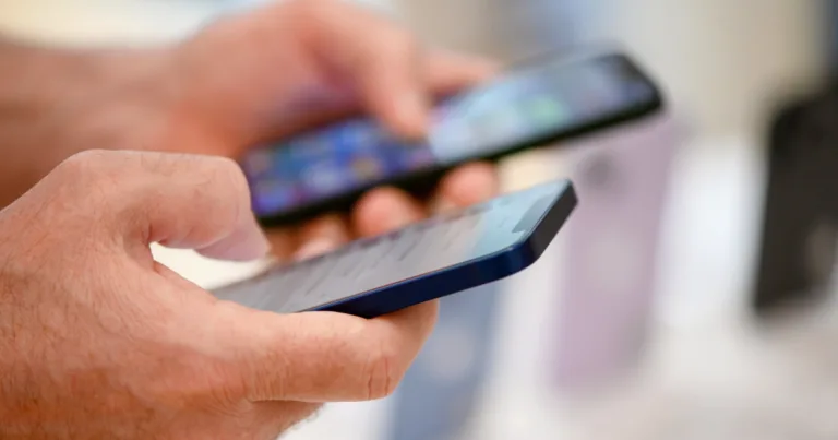 A shopper holds and compares two smartphones in a retail store, with more phones on display in the blurred background.