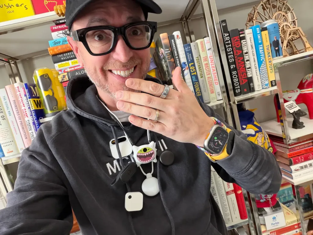 Greg Swan smiling at the camera, wearing thick black-framed glasses and several wearable AI devices on a lanyard around his neck, in front of a bookshelf.