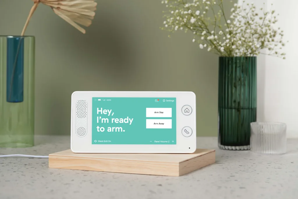 Cove touchscreen security panel on a wooden stand displaying an arming screen with "Arm Stay" and "Arm Away" options, styled on a countertop with decorative vases in the background
