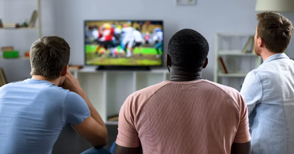 Three friends watch a college football game on TV from a living room couch.