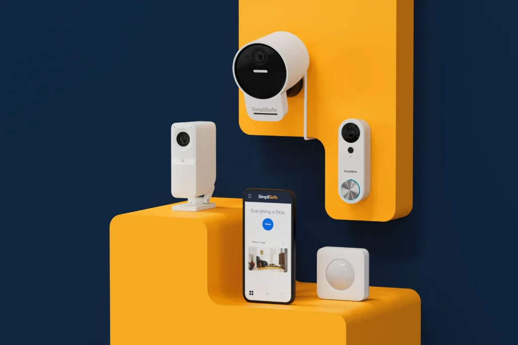 SimpliSafe outdoor camera, indoor camera, video doorbell, motion sensor, and mobile app displayed together on a yellow stepped platform against a dark blue background