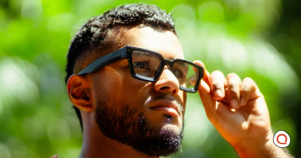 Man outdoors adjusting thick black square-framed glasses while looking into the distance.