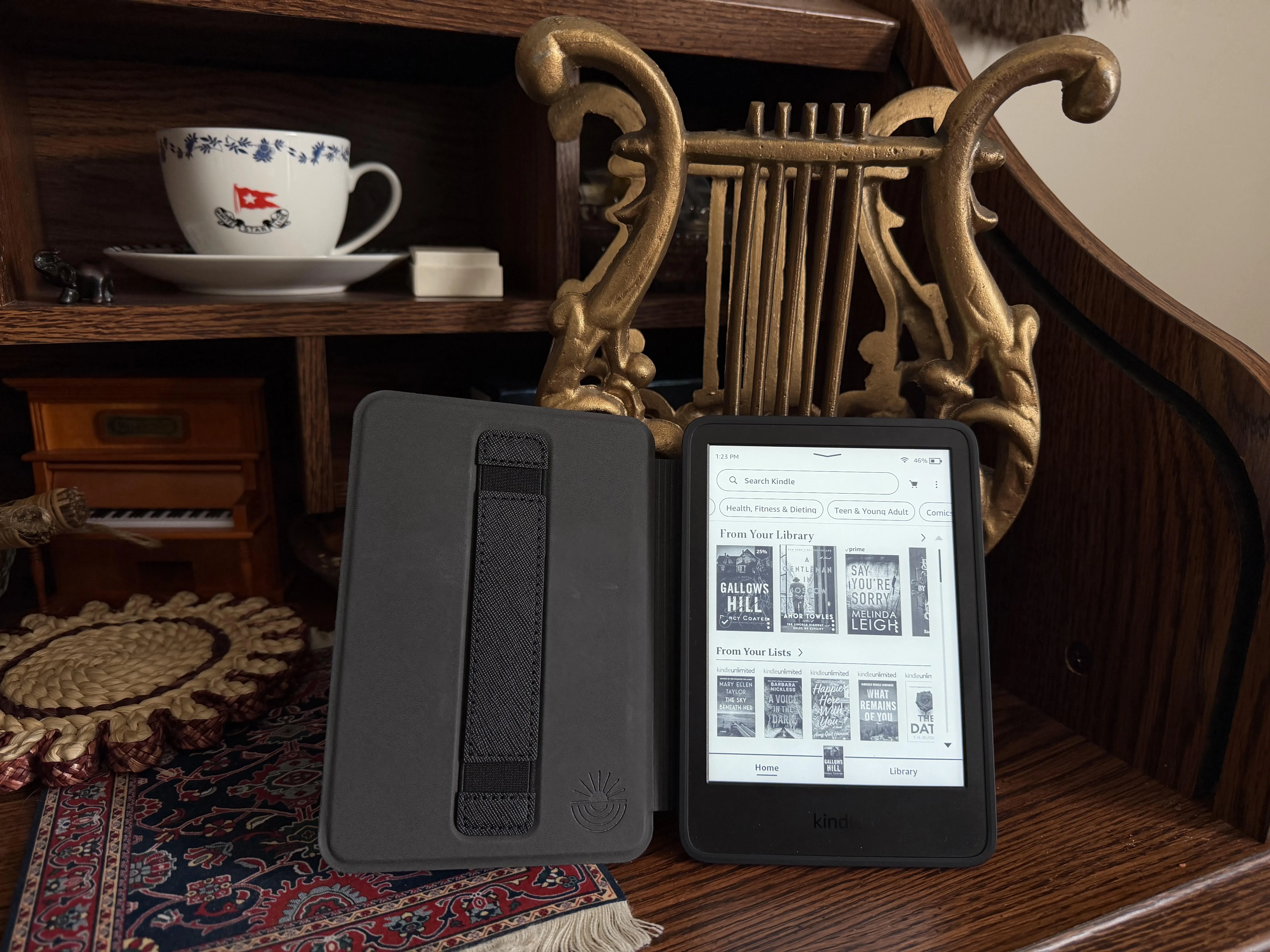 A Kindle Paperwhite 12 Generation lays open on a desk. The screen shows a library including current reads, books from my list, and a search bar.