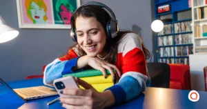 College student smiling at her phone while studying with headphones on, representing an affordable phone and internet combo for student budgets.