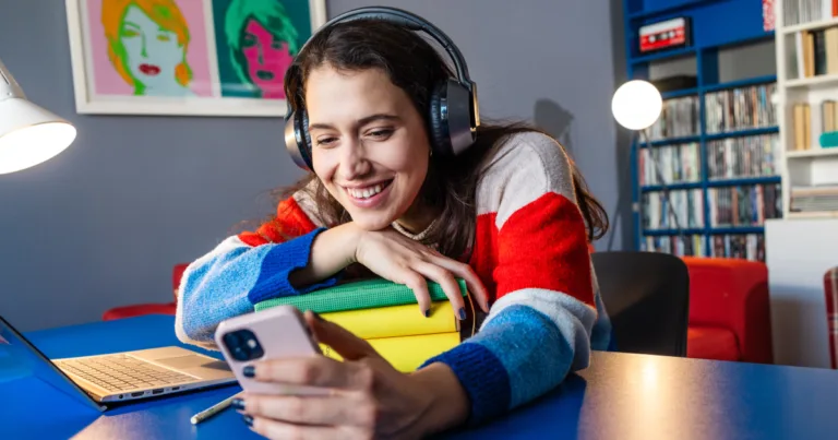 College student smiling at her phone while studying with headphones on, representing an affordable phone and internet combo for student budgets.