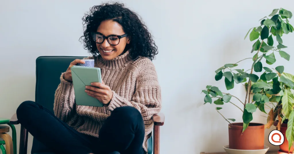 Woman wearing glasses and a beige sweater smiling while holding a tablet device in her hands, sitting in a chair with a potted plant in the background.