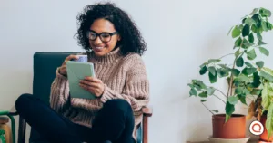 Woman wearing glasses and a beige sweater smiling while holding a tablet device in her hands, sitting in a chair with a potted plant in the background.
