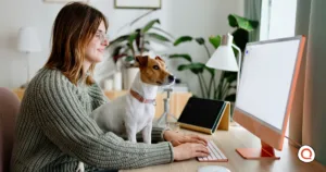 Woman wearing glasses and a green cardigan smiling while working at a wooden desk on a computer, with a brown and white small dog sitting beside her, in a bright home office with plants and natural lighting.