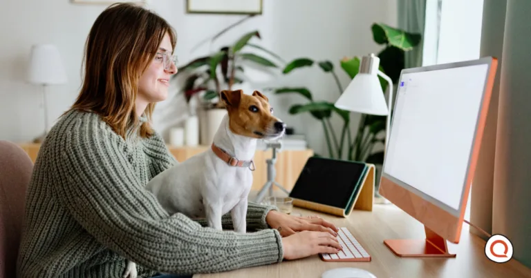 Woman wearing glasses and a green cardigan smiling while working at a wooden desk on a computer, with a brown and white small dog sitting beside her, in a bright home office with plants and natural lighting.