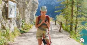 Young woman wearing a climbing helmet and safety harness checks her satellite messenger device while hiking along a mountain trail overlooking a turquoise alpine lake