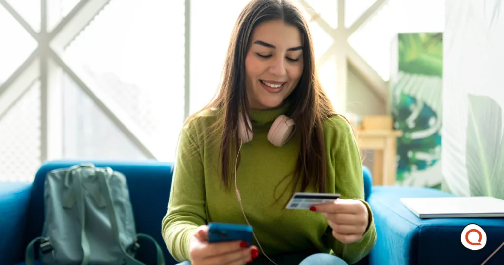 Young woman smiling while making an online payment on her smartphone and holding a credit card, sitting on a blue couch with headphones around her neck