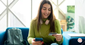 Young woman smiling while making an online payment on her smartphone and holding a credit card, sitting on a blue couch with headphones around her neck