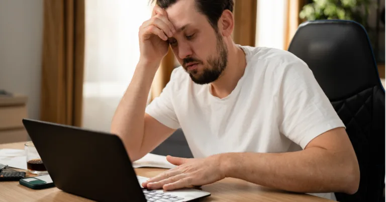 Frustrated man rubbing his forehead while staring at a slow-loading laptop, representing the signs of internet throttling from an ISP.