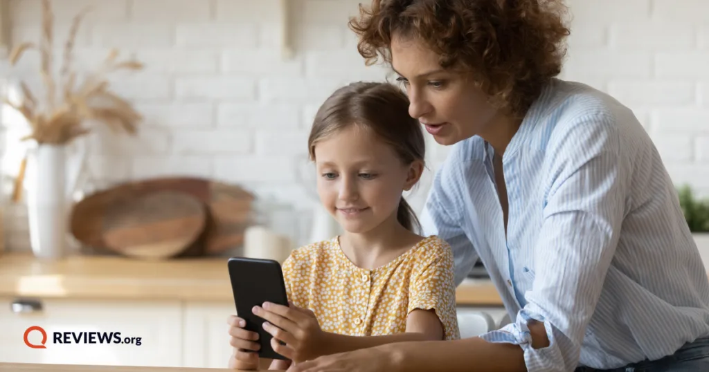 A mother guides her young daughter as she uses a smartphone, showing supervised early phone use at home.