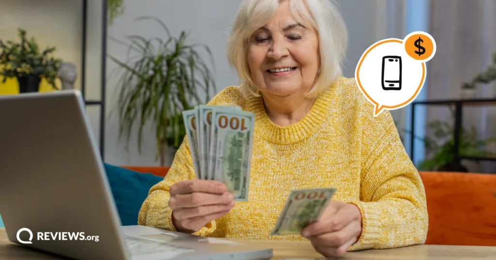 A smiling older woman counts $100 bills at her laptop, showing the monthly savings she could get by switching to a cheaper MVNO phone plan.