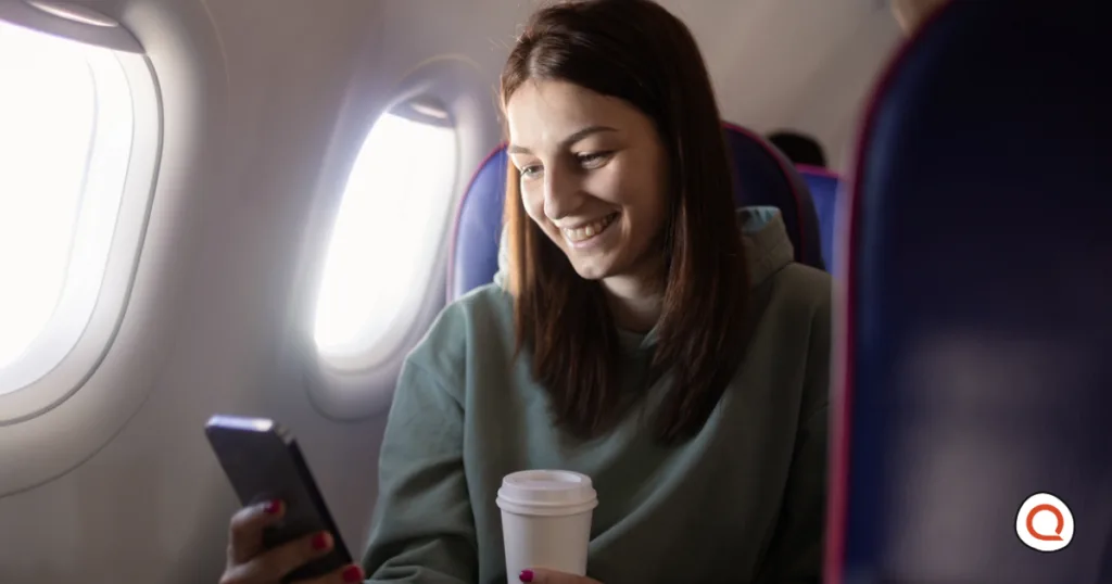 Smiling woman in a green hoodie using her phone in airplane mode while holding a coffee cup in a window seat on an airplane.