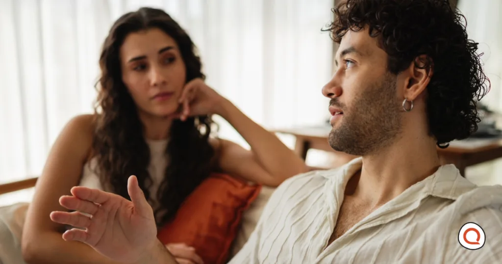 Couple sitting on a couch discussing how to split bills. The man in a cream linen shirt gestures as he talks, and the woman with long curly hair rests her head on her hand and listens.