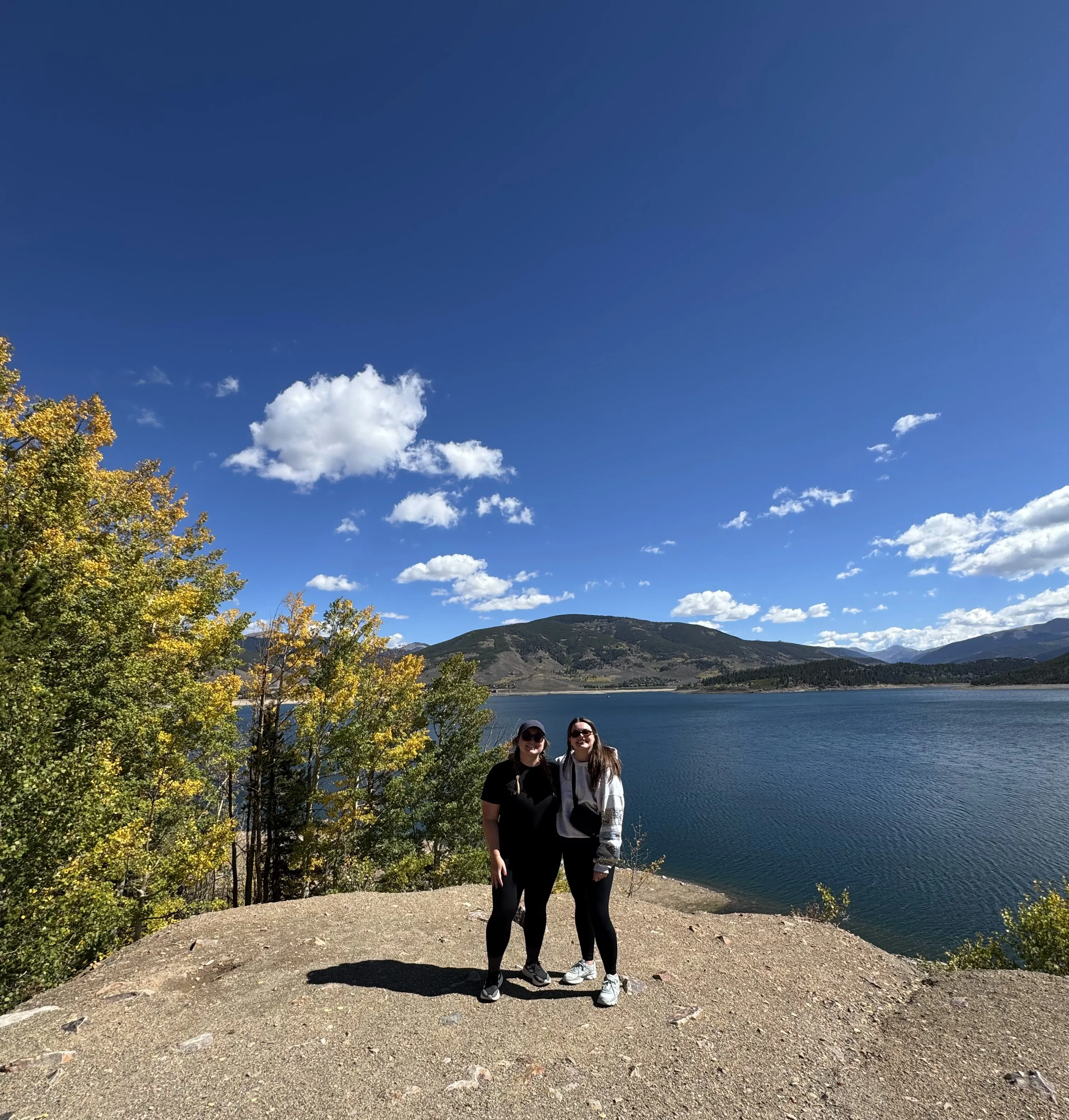 Two friends hiking in the fall leaves in front of a lake.